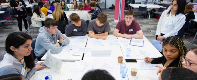 A group of teenagers of mixed genders, sitting around a table covered in paperwork. They are in discussions mimicking climate negotiations at a Mock COP event.